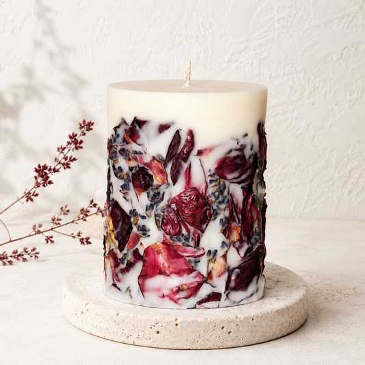 Ivory pillar candle decorated with burgundy rose petals and lavender flowers, displayed on a round travertine stand against a light textured wall.