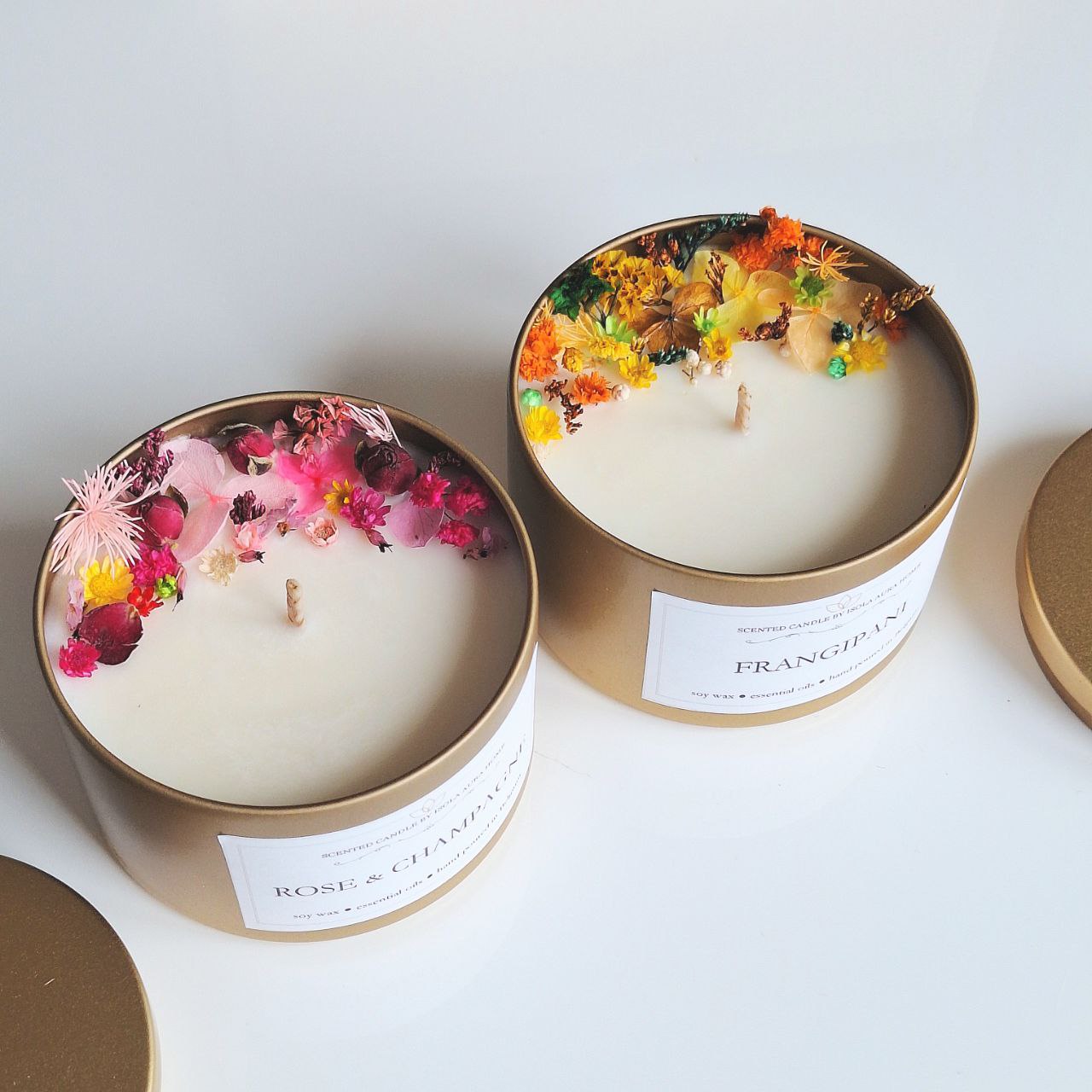 Two designer floral soy wax candles in gold tins with pink and yellow botanical decoration, handmade by Isola Aura Home in Belgium